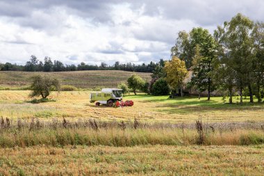 Modern tarımsal hasat makinesi, bulutlu gökyüzünün altındaki ağaçlarla çevrili geleneksel bir çiftlik evinin yakınındaki kırsal alanda çalışıyor..