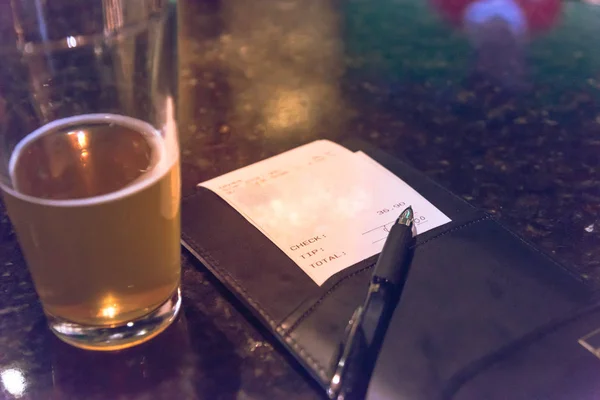 Vintage open pint glass beer next to leather bill holder with restaurant check and pen. Close-up, soft focus receipt total amount on marble table at bar, pub counter. Customer payment for beverage