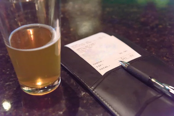 Top view open pint glass beer next to leather bill holder with restaurant check and pen. Close-up, soft focus receipt total amount on marble table at bar, pub counter. Customer payment for beverage