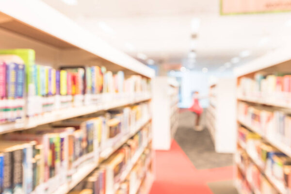 Blurred abstract background of public library interior with bookshelf aisle full of textbooks, literature and magazines. Female student is selecting books from bookshelf. Self-study education concept.