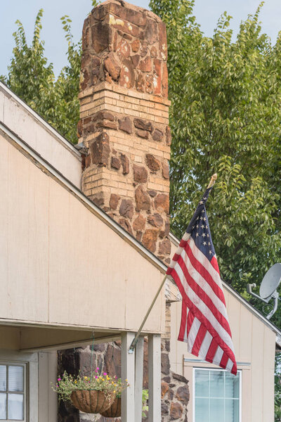 Entrance, front porch of typical one story house proudly displaying American flag. Hanging flower pots near chimney