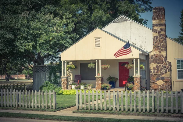 Vintage modest house with chimney in historic downtown district of Irving, Texas, USA. Classic wooden fence with well-groomed landscape, haning flower pots, big tree and proudly American flag waving