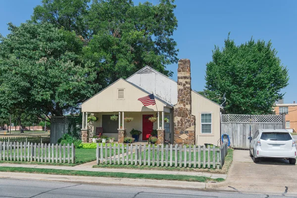Modest house with chimney in historic downtown district of Irving, Texas, USA. Classic wooden fence with well-groomed landscape, haning flower pots, big tree and proudly American flag waving