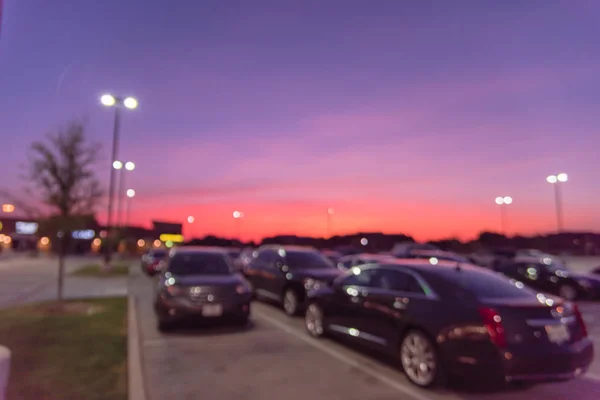 Abstract blurred exterior of modern commercial strip in Irving, Texas, US at sunset. Shopping center row of cars in outdoor uncovered parking lots, bokeh of retail store, light poles in background