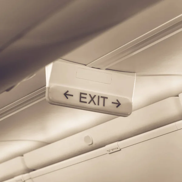 Vintage tone emergency exit sign with red arrow on ceiling of airplane. Illuminated evacuation symbol underneath the overhead compartments. Shining signboard show escape of emergency, urgency concept