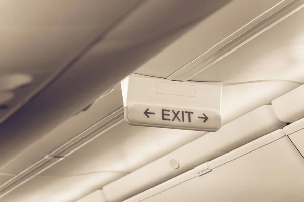 Vintage tone emergency exit sign with red arrow on ceiling of airplane. Illuminated evacuation symbol underneath the overhead compartments. Shining signboard show escape of emergency, urgency concept