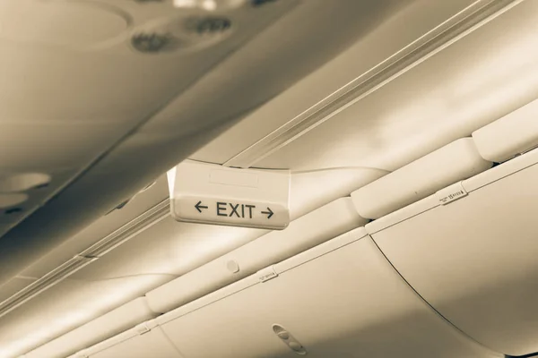 Vintage tone emergency exit sign with red arrow on ceiling of airplane. Illuminated evacuation symbol underneath the overhead compartments. Shining signboard show escape of emergency, urgency concept