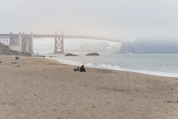 Rear view people sitting on the shore enjoy Baker Beach during foggy summer day. Golden Gate bridge in the background