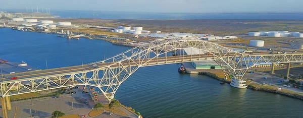 Panorama aerial view of Corpus Christi Harbor Bridge with row of oil tanks and wind turbines farm in distance. A through arch bridge crosses the Corpus Christi Ship Channel