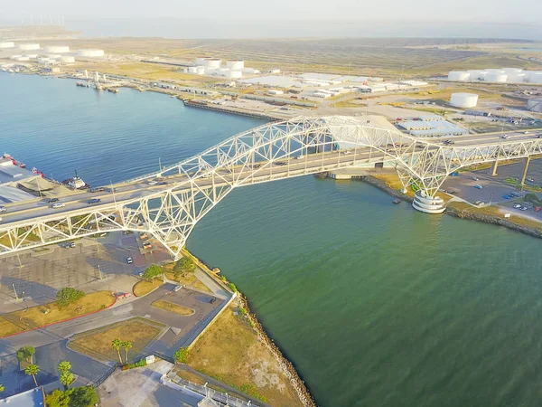 Aerial view of Corpus Christi Harbor Bridge with row of oil tanks and wind turbines farm in distance. A through arch bridge crosses the Corpus Christi Ship Channel