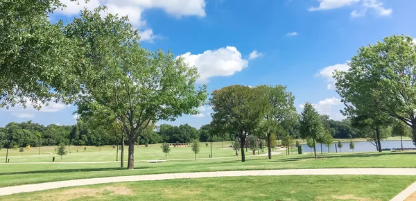 Panorama view green and clean lakeside park with pathway trail system in Coppell, Texas, USA. Grassy lawn park with mature trees under sunny summer cloud blue sky