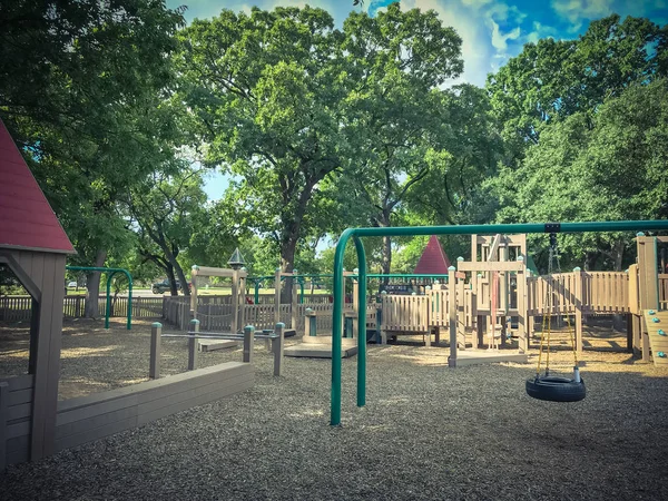 Tire and chair swing set at public wooden children playground under the lush of larger trees in Coppell, Texas, USA. Play set structure surrounded by summer leaves green and cloud blue sky