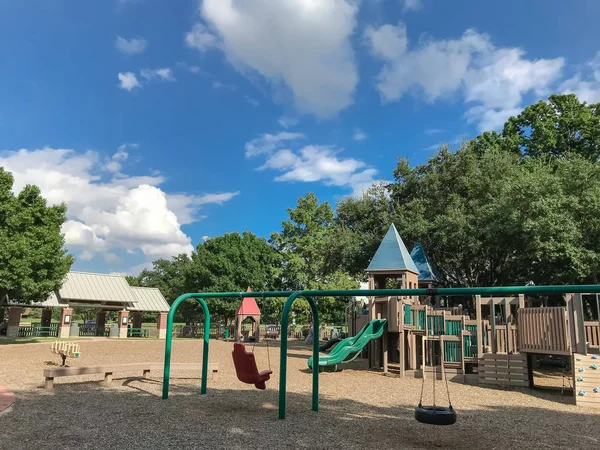 Tire and chair swing set at public wooden children playground under the lush of larger trees in Coppell, Texas, USA. Play set structure surrounded by summer leaves green and cloud blue sky