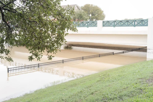 Flooded river pathway trail under bridge in suburban Dallas Fort Worth area, Texas, USA. Metal fence panel stands underwater, pedestrian road drowned by high water overflow