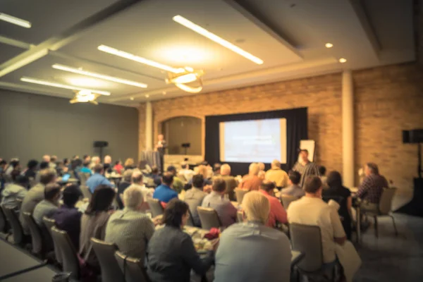 Blurred business seminar meeting with LED projector screen and speaker speech on stage. Defocused rear view audience in conference hall room, listening talk show in USA. Education, business concept