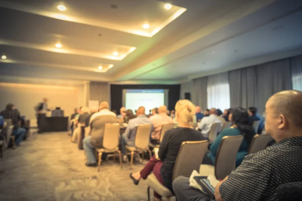 Vintage tone blurred business seminar meeting with LED projector screen, speaker speech on stage. Defocused rear view audience in conference hall room, listening talk show. Education, business concept