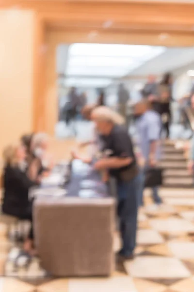 Blurred side view diverse participants at registration check-in workshop table. Multiethnic people check-in at hotel lobby near staircase and received instruction, event package from support staff