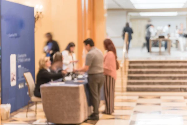 Motion blurred participants check-in at registration table of workshop in hotel lobby near marble grand staircase. Multiethnic people sign-up and received instruction, event package from support staff