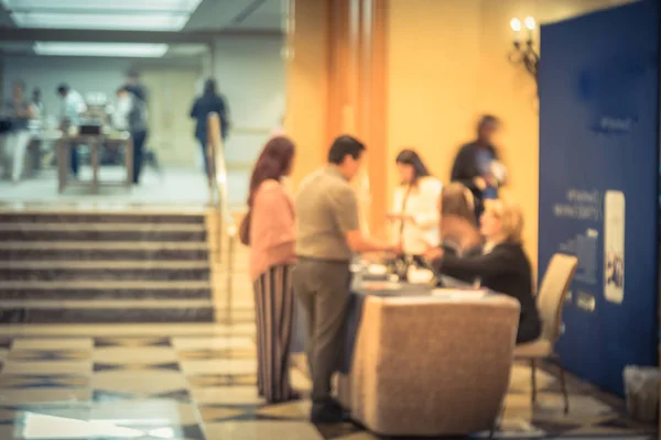 Motion blurred participants check-in at registration table of workshop in hotel lobby near marble grand staircase. Multiethnic people sign-up and received instruction, event package from support staff