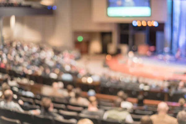 Blurred abstract Christian people inside the church listening to preacher speaking. Defocused back view audience on row of raised sitting chairs looking at stage with large video projector screens