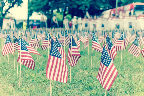 Filtered image lawn American flags with blurry row of people carry fallen soldiers banners parade