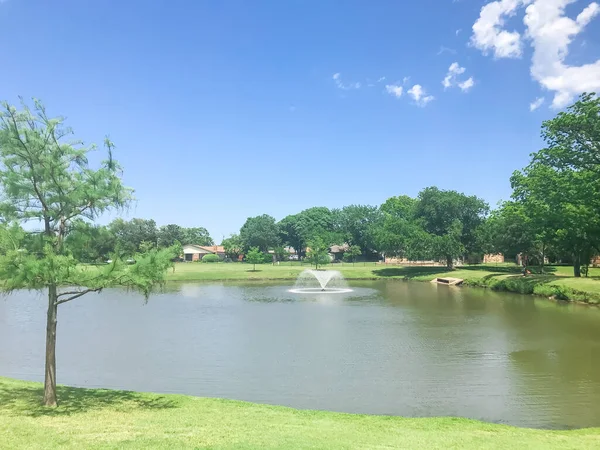 Working water fountain at residential park with clear pond and green tall trees in Coppell, Texas, USA