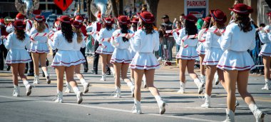 Panorama manzaralı beyaz etekli değnek çeviriciler, bayram geçit töreninde büyük pirinç enstrümanlı bandoyu takip ediyor. Kırmızı şapkalar, çizmeler, metal barikatlardan izlenen kalabalık gibi sezonluk yetenekler ekliyor, Dallas. ABD