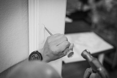 Cesur adam kapı çerçevesine tornavida ve çekiç kullanarak grev levhası yerleştiriyor. Yönlendirilmiş işaret, dikdörtgen aralıkları oyuyor. Arka planda bulanık mobilyalar ve tamir sırasında evin içi görünüyor. ABD
