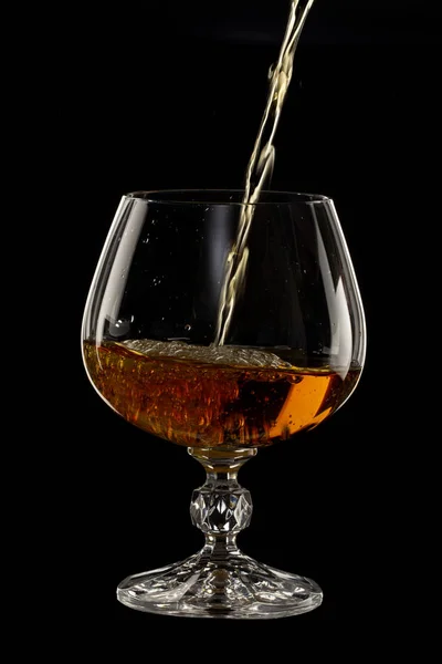 Cognac is poured into a glass, a spray of a drink, on a black background close up