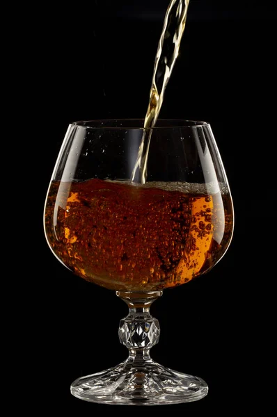 Cognac is poured into a glass, a spray of a drink, on a black background close up