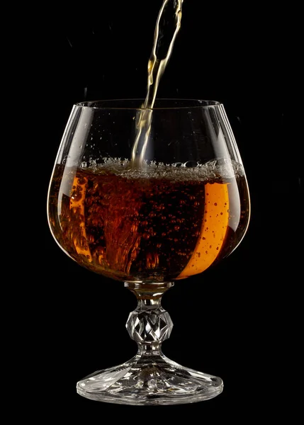 Cognac is poured into a glass, a spray of a drink, on a black background close up