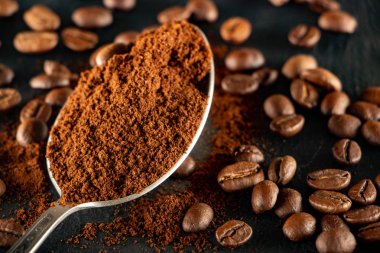 Öğütülmüş kahveli metal bir kaşık, kahve tohumlarının yayıldığı bir kayrak tepsisi üzerinde yer alır..