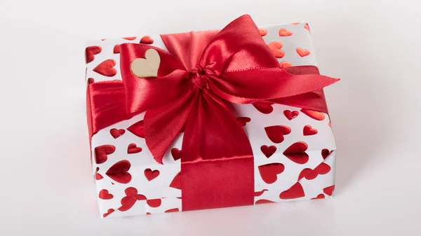 White box with a birthday surprise, beloved girl on a white background. Wrapped in paper with a heart-shaped texture, and tied with a red ribbon with a bow.