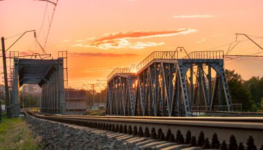 Gün batımında yeşil kıyıları olan küçük bir nehir üzerindeki eski metal demiryolu köprüsü..