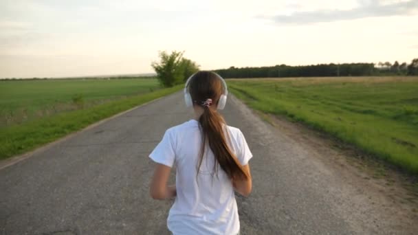 Chica con auriculares de música corre a lo largo de la carretera. hermosa chica haciendo ejercicios deportivos, cámara lenta — Vídeo de stock