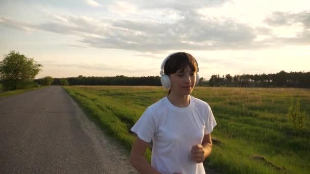 Chica joven en auriculares de música que se ejecutan en el entrenamiento en carretera, estilo de vida deportivo, cámara lenta — Vídeo de stock