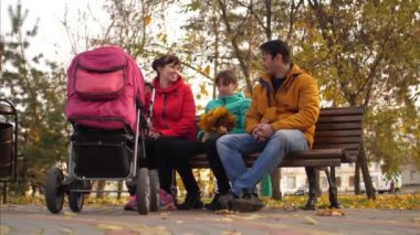 Anne ve baba çocukları ile sonbahar bir parkta bankta bebek arabası ile oturup konuşmak.