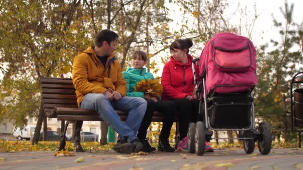 Женатая пара с детьми сидят на скамейке в осеннем парке с коляской и разговаривают . — стоковое видео