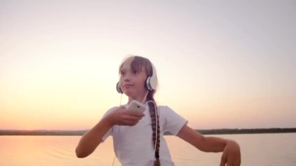 Teenage girl dances in headphones with phone on against sunset. — Stock Video