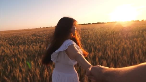 La fille court à travers le champ avec du blé tenant une main d'homme dans l'éblouissement du soleil couchant et rit. Mouvement lent . — Video