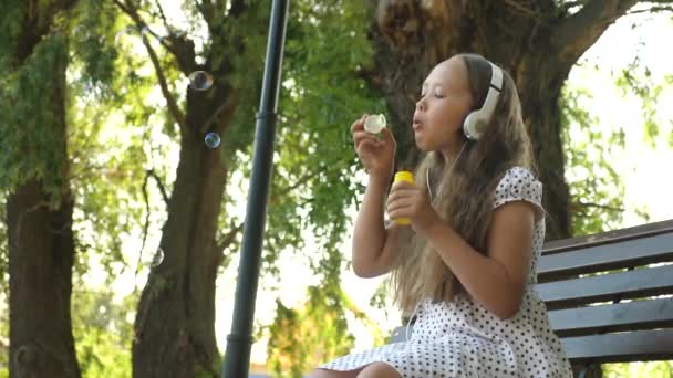 Dospívající dívka v sluchátka sedí na lavičce a fouká mýdlové bubliny a úsměvy. — Stock video