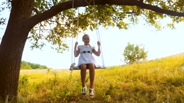 Glad tjej med långt hår gungor på gunga under sommaren ek i en vit klänning och skrattar. — Stockvideo