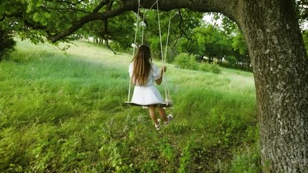 Vrouwtje met lange haren in een witte jurk swingen op een schommel onder een zomer eiken boom in het warme seizoen. Slow motion. — Stockvideo