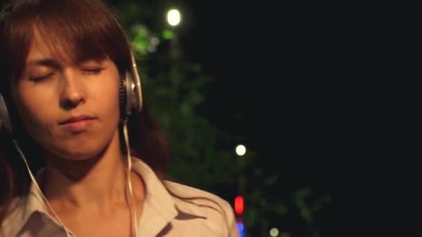 Chica en auriculares con pelo largo y teléfono táctil bailando sonriendo y disfrutando de la música por la noche en el parque. Primer plano — Vídeos de Stock