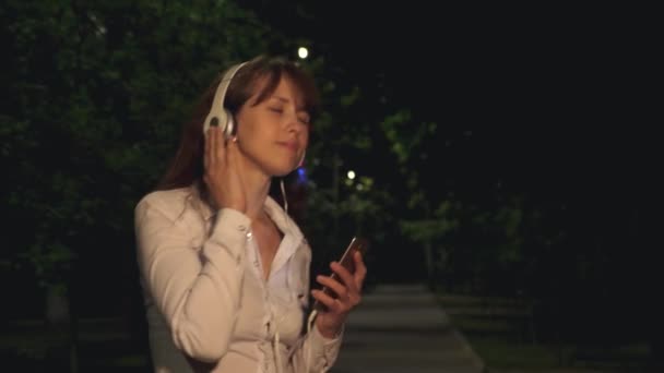 Chica en auriculares y teléfono táctil agitando el pelo largo y bailando sonriendo y recibiendo zumbido de la música por la noche en el parque — Vídeos de Stock