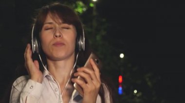 Kız kulaklık ve uzun saç şarkı ve dans müzik gece parkta gülümseyen ve kayfuya sallayarak dokunmatik telefon. Yakın çekim