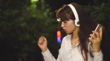 Kız kulaklık ve uzun saç şarkı ve dans müzik gece parkta gülümseyen ve kayfuya sallayarak dokunmatik telefon. Yakın çekim
