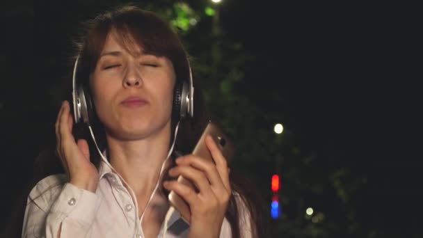 Chica en auriculares y teléfono táctil agitando el pelo largo cantando y bailando sonriendo y kayfuya de la música por la noche en el parque. Primer plano — Vídeos de Stock