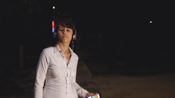 Giovane ragazza in cuffia con touch phone balla e canta sorridendo e ricevendo una scarica di adrenalina dalla musica di notte nel parco cittadino — Video Stock