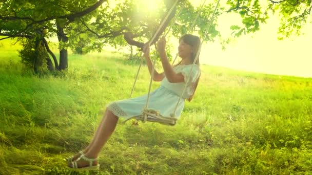 Jong meisje swingt op een schommel in de schittering van een gouden zonsondergang en glimlacht. Slow motion. — Stockvideo